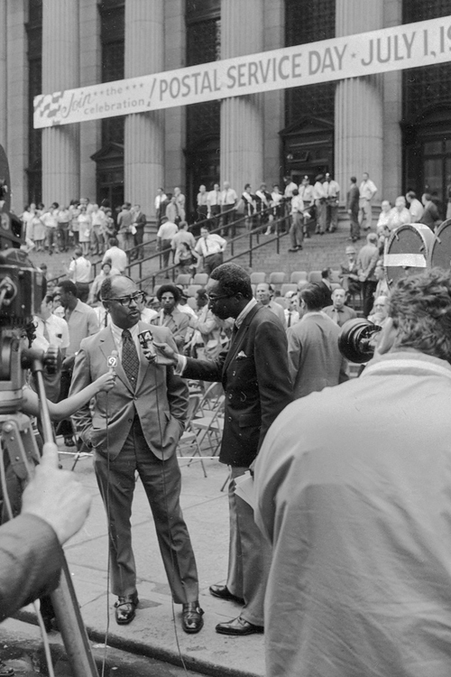 A black-and-white image of a Black man in a business suit standing on a New York City sidewalk talking to TV news crews