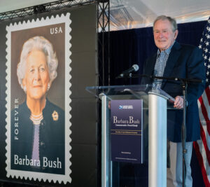 Former president George W. Bush, wearing a suit jacket, stands at a lectern near a Barbara Bush stamp poster