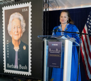 A woman in a business suit stands at a lectern near a Barbara Bush stamp poster