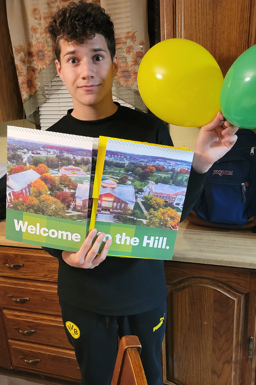 A young man stands in a kitchen and holds a folder that reads “Welcome to the Hill.”