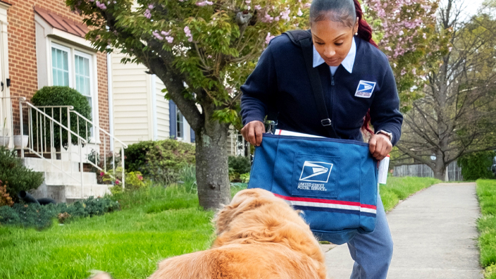 A woman in a postal uniform crouches in a defensive posture, holding her mail satchel as a shield as a dog approaches