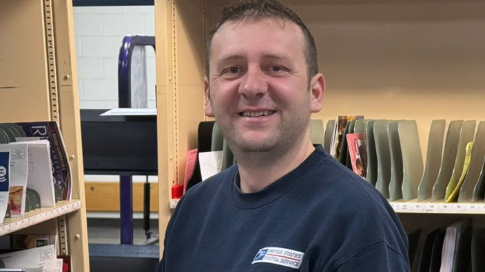 A smiling man in a long-sleeve Postal Service shirt stands near a letter sorting shelf