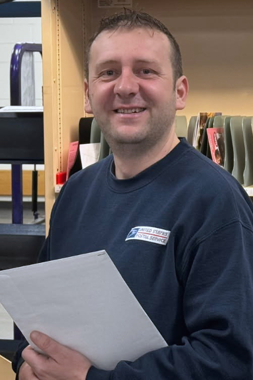 A smiling man in a long-sleeve Postal Service shirt stands near a letter sorting shelf