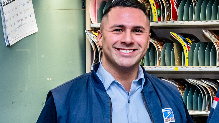 A smiling man in a sharp USPS uniform stands in a Post Office workroom