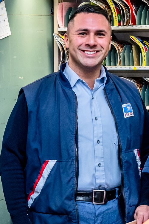 A smiling man in a sharp USPS uniform stands in a Post Office workroom