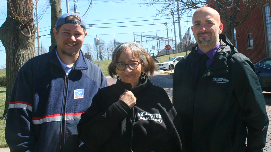 Two men in postal uniforms stand next to a woman on a sidewalk