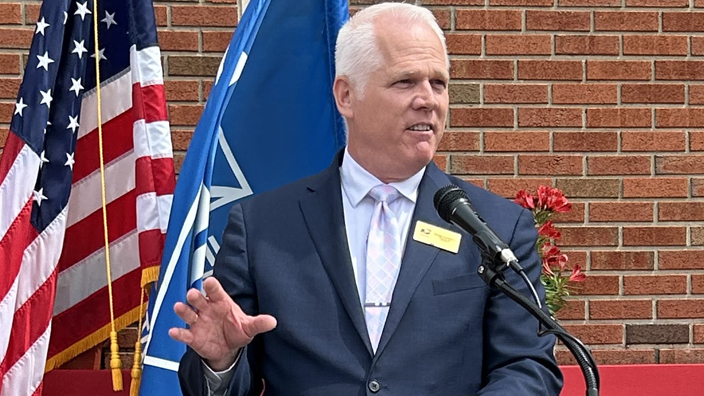 A man in a business suit stands at a lectern with the U.S. and other flags on the podium behind him