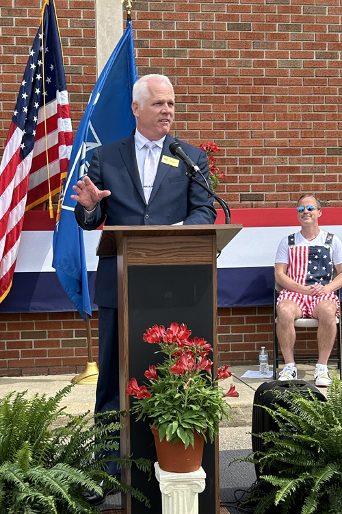 A man in a business suit stands at a lectern with the U.S. and other flags on the podium behind him