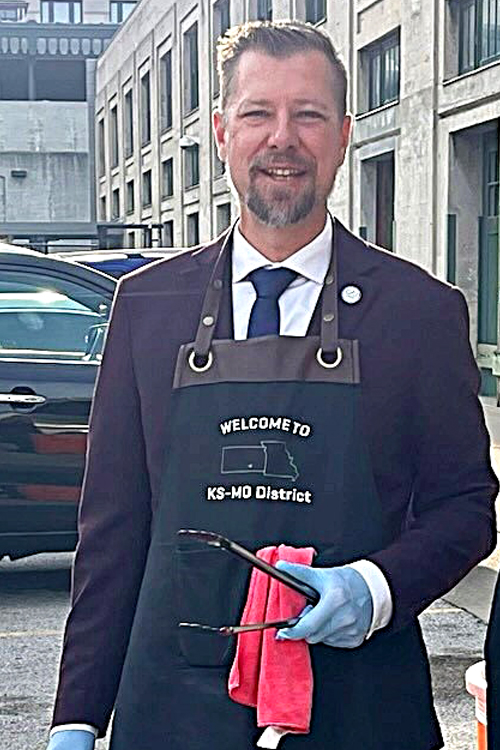 A man wearing an apron over a business suit stands next to a grill in a parking lot