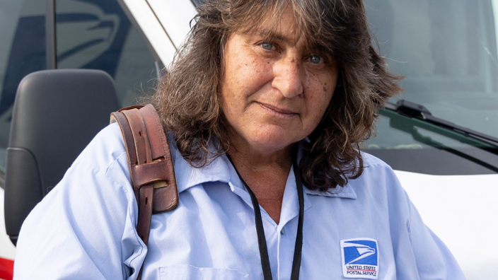 A woman in a postal uniform stands by a mail delivery truck and points to a spot on her right hand