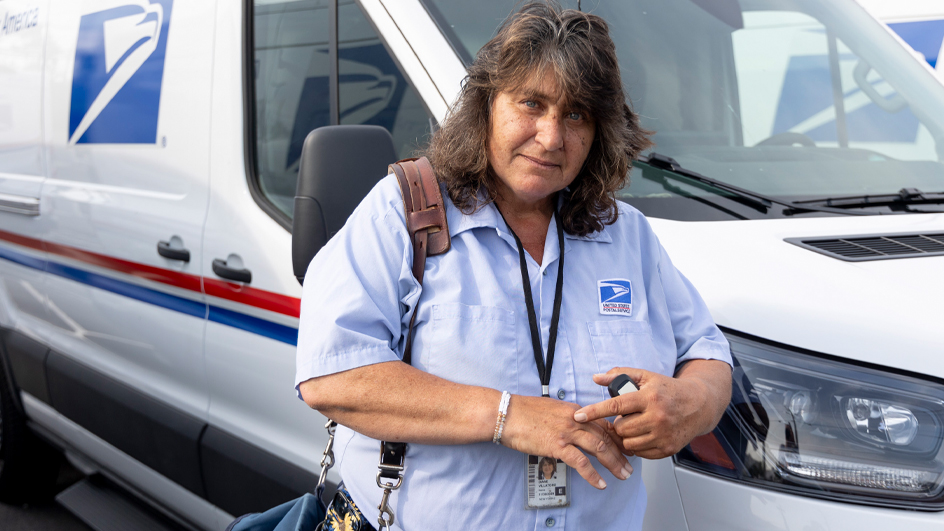 A woman in a postal uniform stands by a mail delivery truck and points to a spot on her right hand