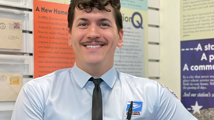 A man dressed in a dapper postal uniform, complete with tie, smiles in a Post Office lobby decrated with historical artifacts and posters