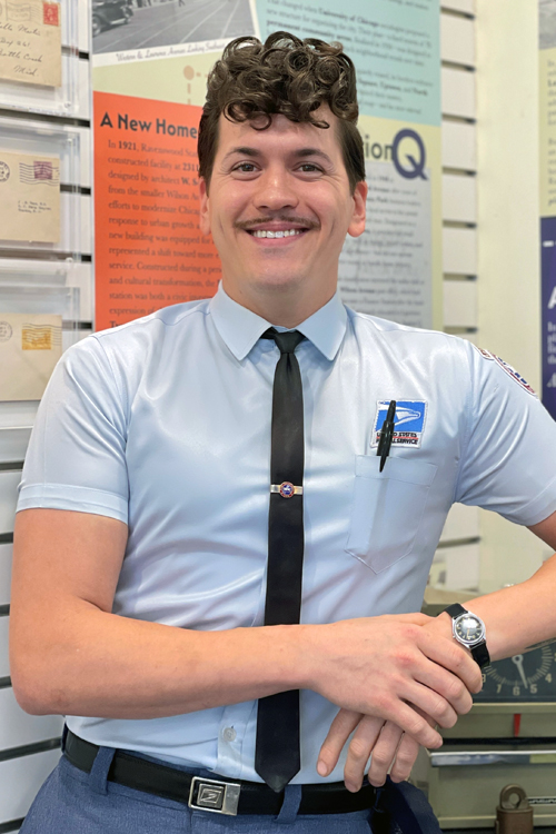 A man dressed in a dapper postal uniform, complete with tie, smiles in a Post Office lobby decrated with historical artifacts and posters