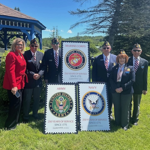 A woman in a red jacket and five people in blue jackets wearing military hats and pins stand around posters of stamps.