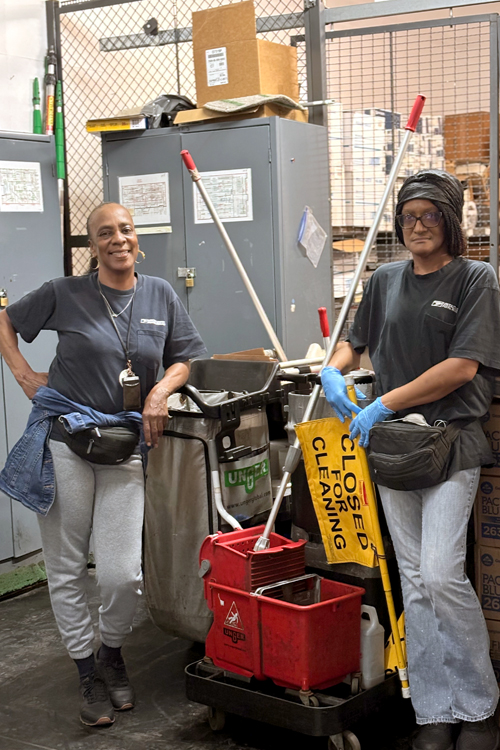 Two women smile and stand among cleaning carts and equipment