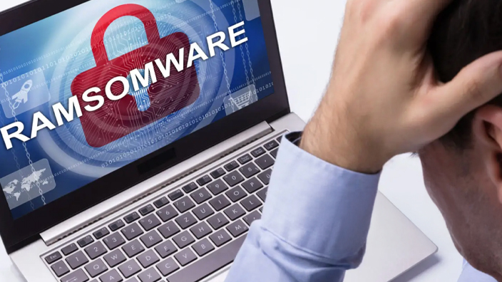 A man seated at his desk buries his head in his hands. Near him, a computer monitor displays the word “ransomware.”