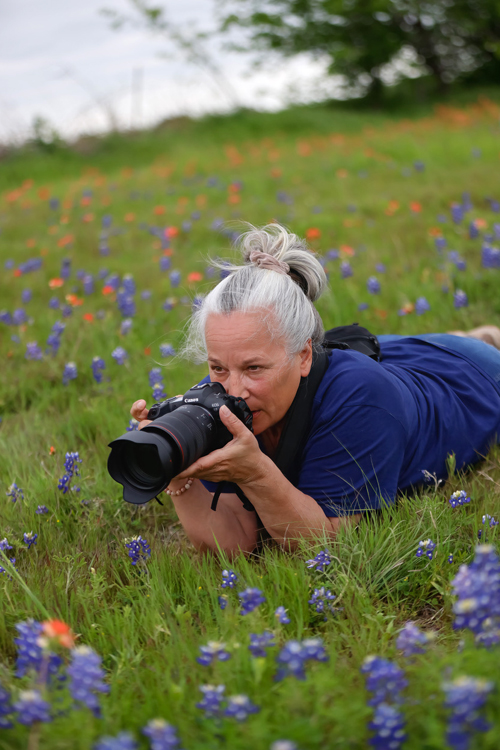 A woman lies on her stomach in a lush field and peers over a long-lens camera