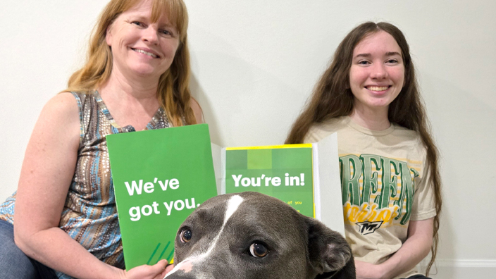 A smiling young woman holds a brochure while seated next to a woman and a dog