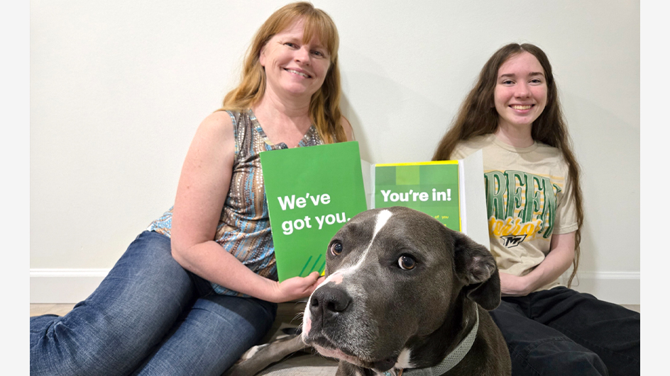 A smiling young woman holds a brochure while seated next to a woman and a dog