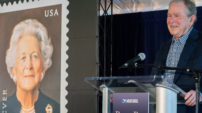 Former president George W. Bush stands at a lectern and speaks near a poster of the Barbara Bush stamp