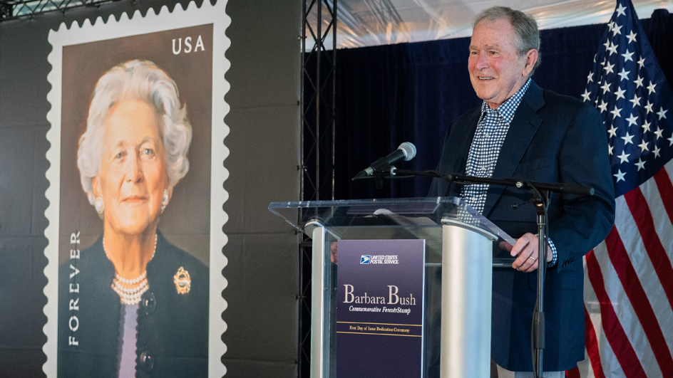 Former president George W. Bush stands at a lectern and speaks near a poster of the Barbara Bush stamp
