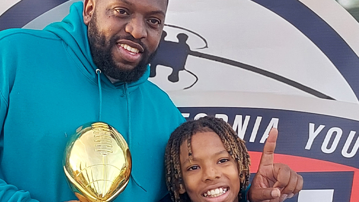 A man in a hoodie smiles and holds a trophy while draping an arm around a smiling boy who is about 10 years old
