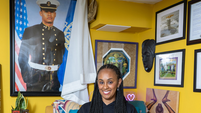 A smiling woman sits in a home office holding a book. Looming on a nearby wall is a large portrait of her late son, dressed in a naval uniform