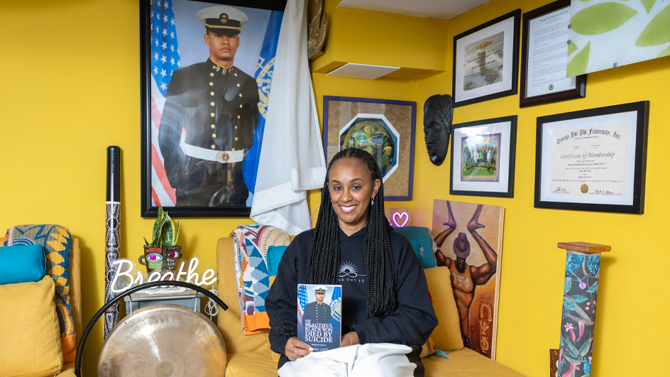 A smiling woman sits in a home office holding a book. Looming on a nearby wall is a large portrait of her late son, dressed in a naval uniform