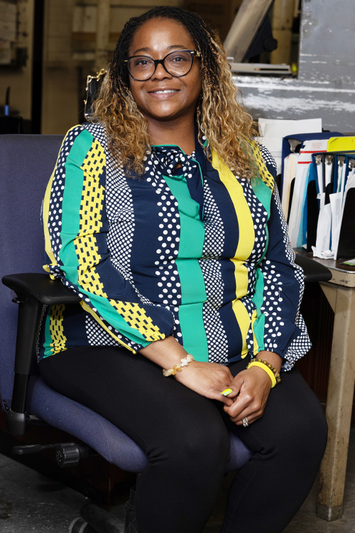 A woman sits in a chair in a Post Office workroom and smiles