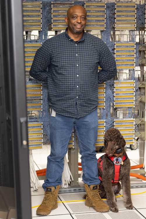 A man wearing jeans, a flannel shirt and boots stands against a bank of technology equipment with a dog at his feet