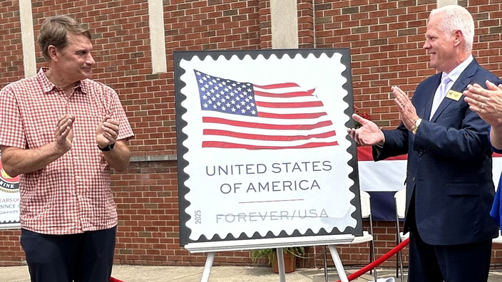 People stand in the parking lot of a brick building and applaud next to a poster of a stamp bearing the U.S. Flag