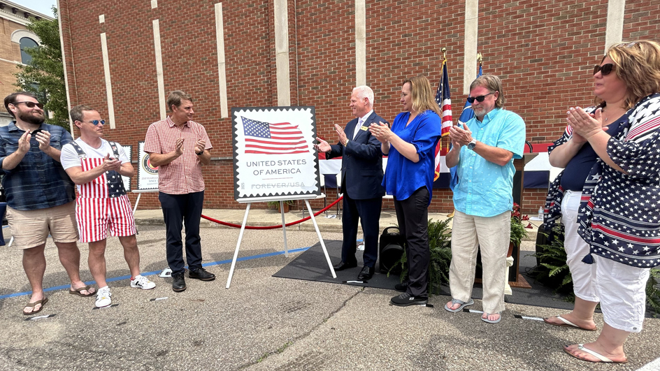 People stand in the parking lot of a brick building and applaud next to a poster of a stamp bearing the U.S. Flag