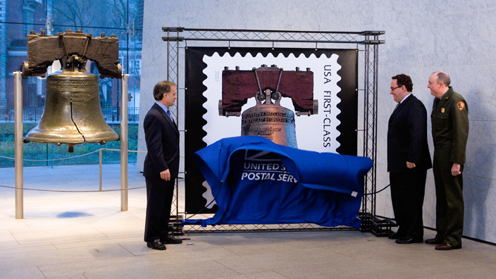 Three men remove a drape covering a stamp image while standing next to the Liberty Bell