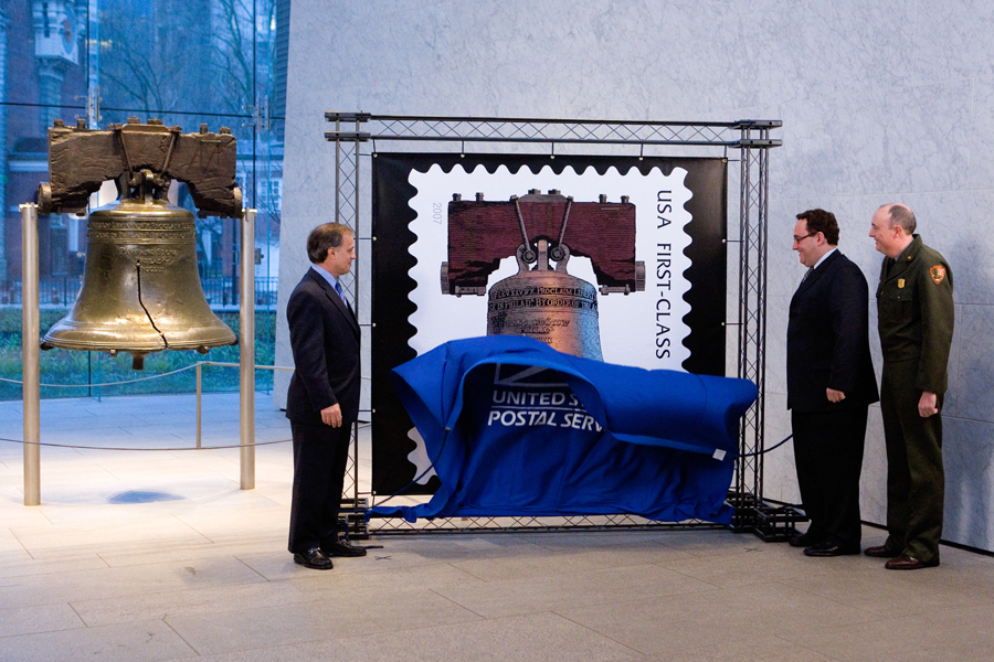 Three men remove a drape covering a stamp image while standing next to the Liberty Bell
