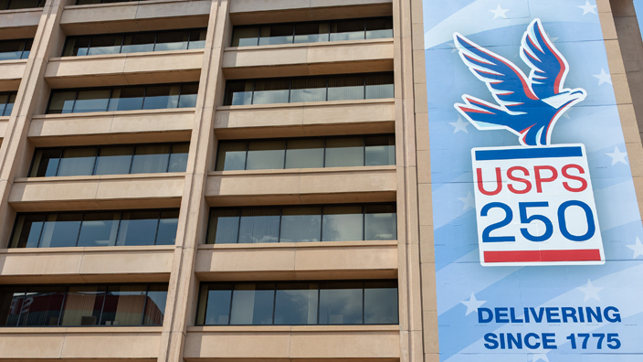 A large banner hanging on the side of an office building. The banner shows an illustration of an eagle about to take wing and the words “USPS 250.”