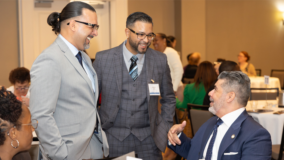 Two young men in business suits stand and speak to an older man seated at a table in a hotel ballroom