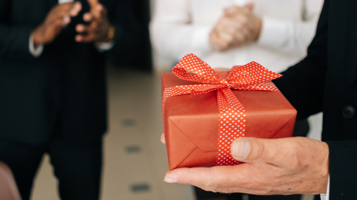 Men in business suits stand in a room, one smiling as another holds a giftwrapped box