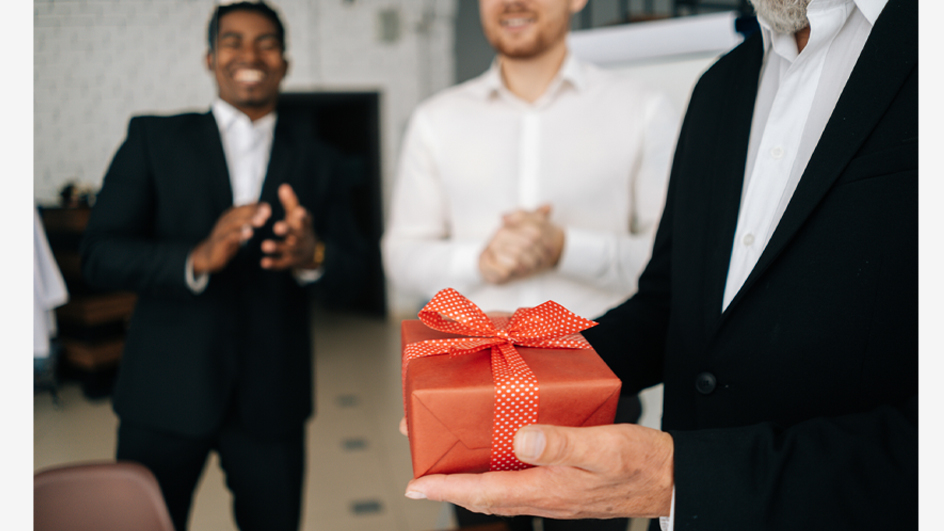 Men in business suits stand in a room, one smiling as another holds a giftwrapped box