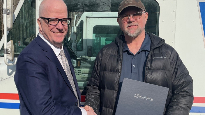 A man in a business suit presents a certificate to a man in a winter coat and jeans in front of a postal delivery vehicle