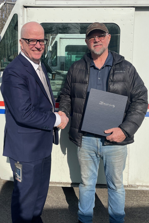 A man in a business suit presents a certificate to a man in a winter coat and jeans in front of a postal delivery vehicle