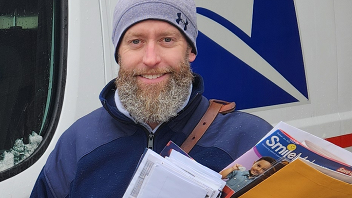A man in a postal uniform smiles as he stands next to a delivery vehicle