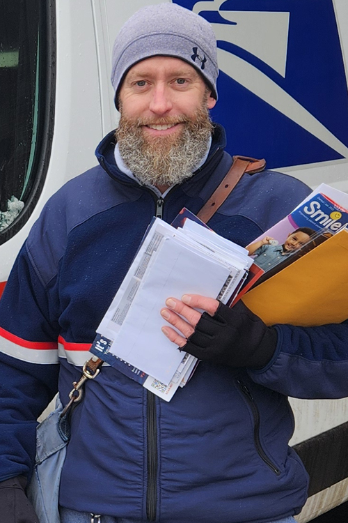 A man in a postal uniform smiles as he stands next to a delivery vehicle