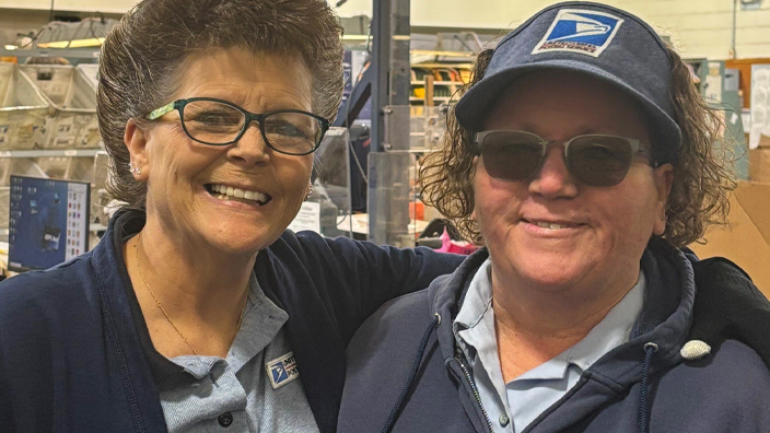 Two women wearing postal uniforms stand next to each other