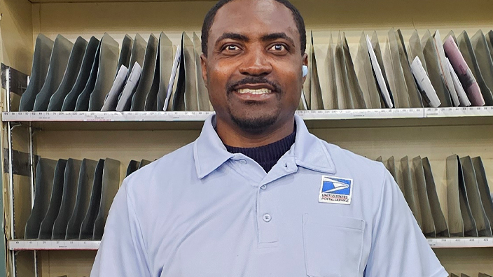 A man in a postal uniform stands in a Post Office workroom, smiling