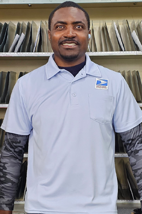 A man in a postal uniform stands in a Post Office workroom, smiling