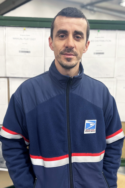 A man in a postal uniform stands in a Post Office workroom