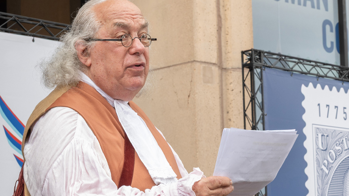 A man dressed as Benjamin Franklin sits on a stage and reads from a sheet of paper in his hand