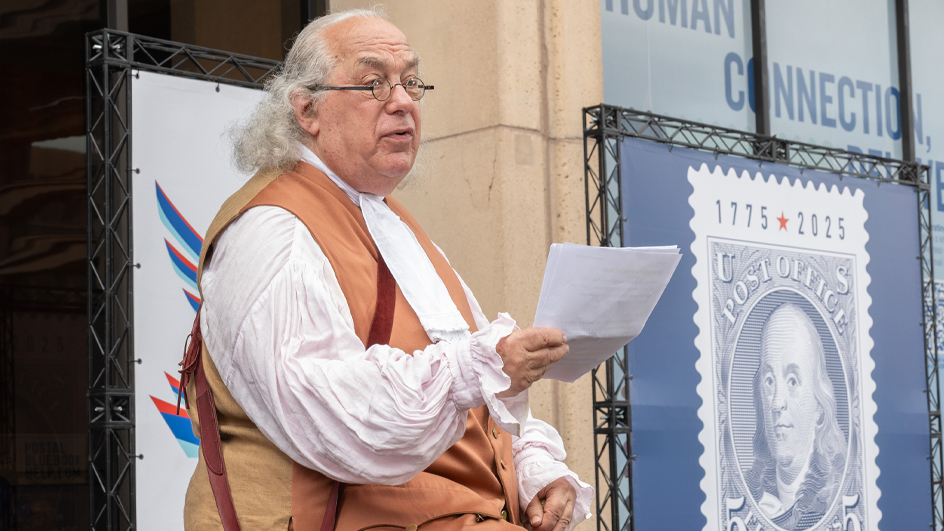 A man dressed as Benjamin Franklin sits on a stage and reads from a sheet of paper in his hand