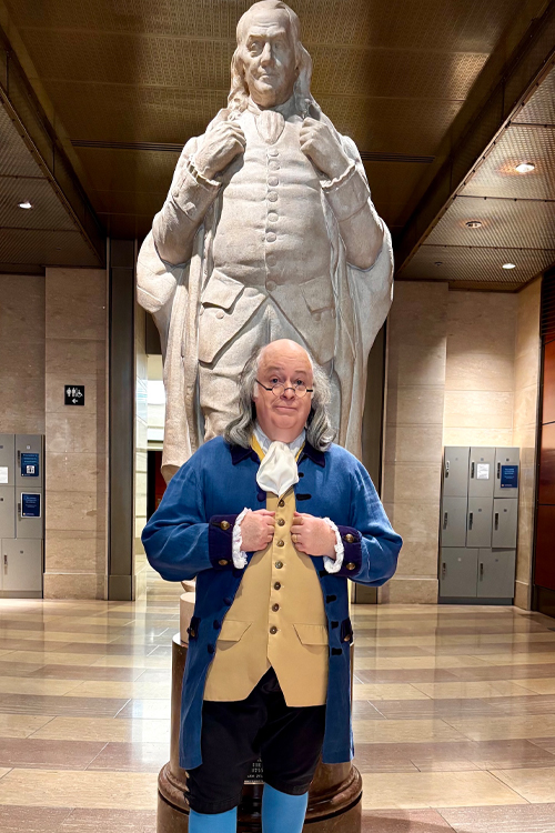 A man dressed as Benjamin Franklin stands in front of a Benjamin Franklin statue