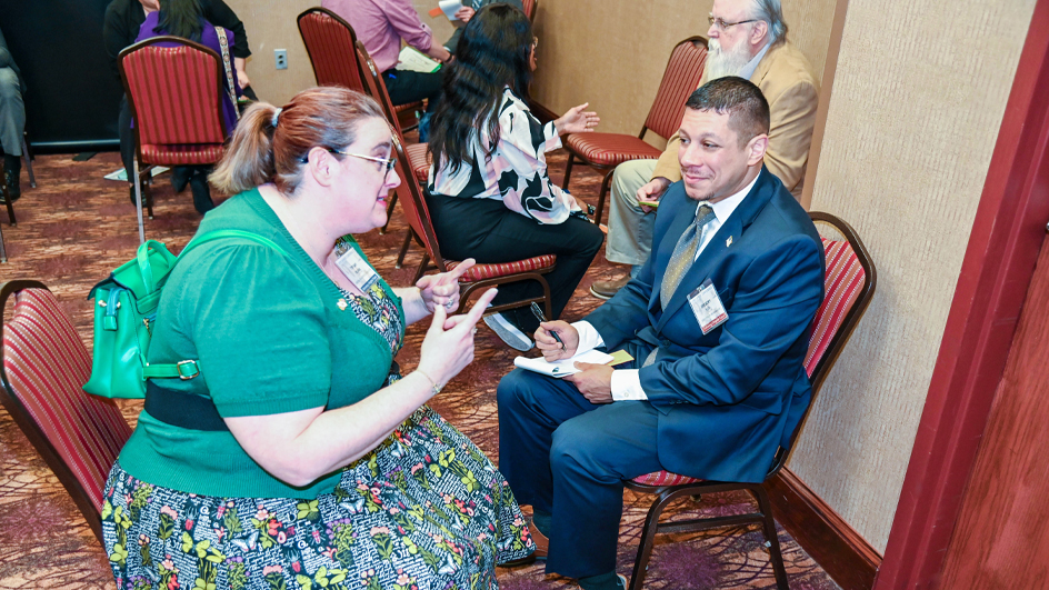 A woman and a man wearing professional business attire sit and smile at a table in a hotel ballroom